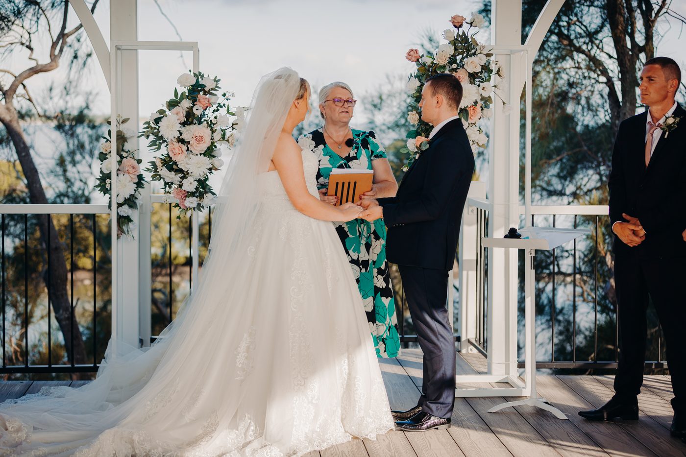 Bride Emily and groom Dylan hold hands facing each other during their wedding ceremony, with the officiant standing between them and a groomsman standing to the side. Valentine's Day wedding photography by She Said Yes.