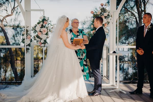 Emily the bride and Dylan the groom hold hands during their wedding ceremony at Sandstone Point Hotel — Pavilion, with floral decorations in the background.