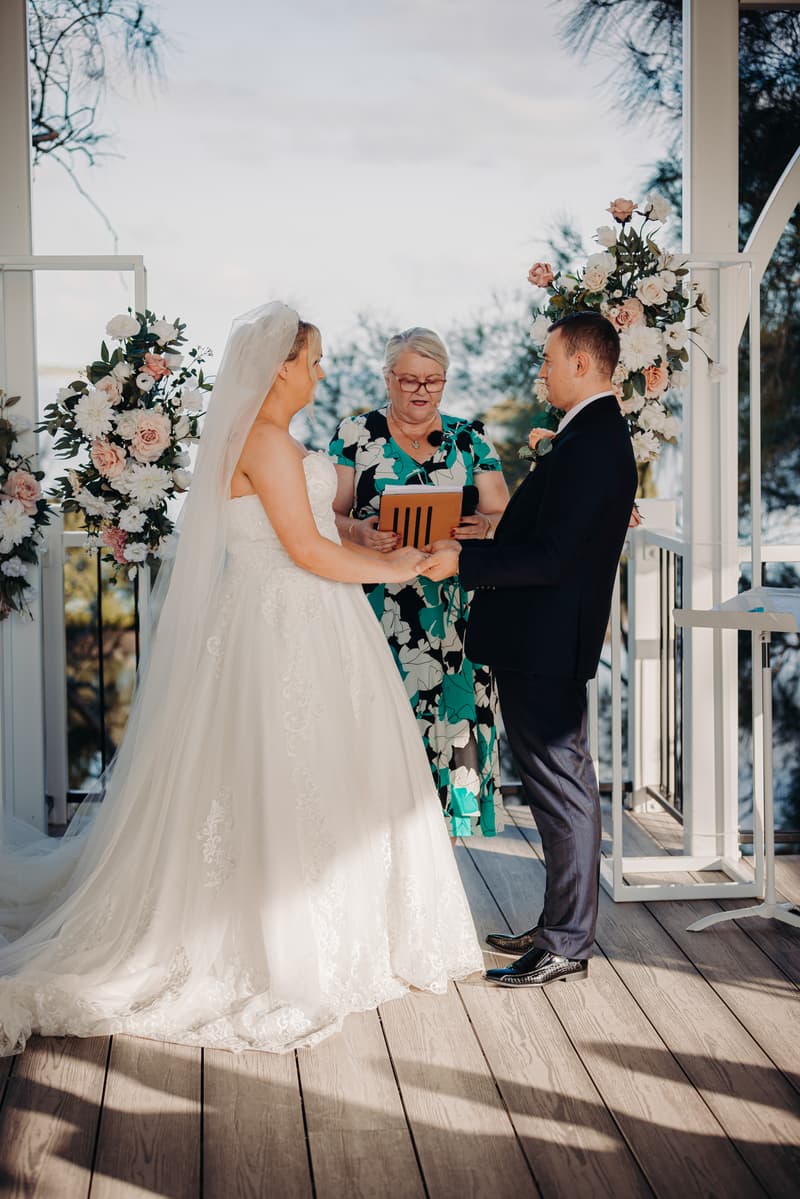 Emily and Dylan hold hands during their wedding ceremony at Sandstone Point Hotel — Pavilion, with the officiant holding a microphone and documents in the background.