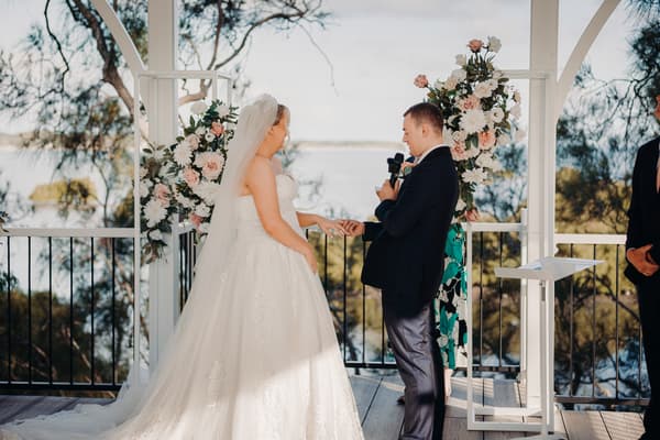 Emily and Dylan exchange rings during their wedding ceremony at Sandstone Point Hotel — Pavilion, with floral arrangements on the altar and a scenic outdoor background.