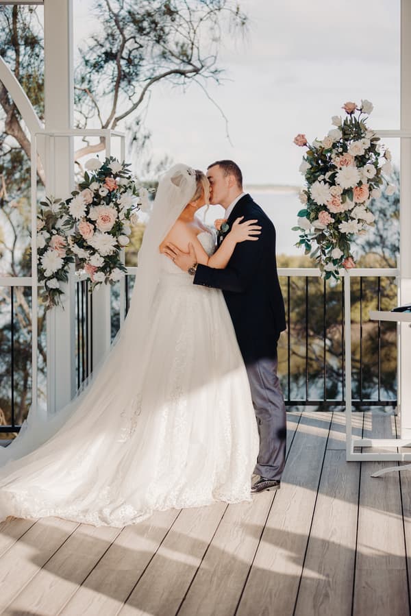 Emily and Dylan share a kiss at the ceremony stage decorated with floral arrangements at Sandstone Point Hotel — Pavilion.