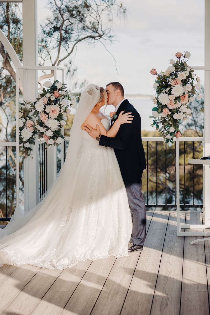 Emily and Dylan share a kiss at the ceremony stage decorated with floral arrangements. May wedding sunset Brisbane by She Said Yes.