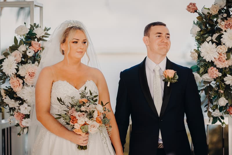 Emily the bride and Dylan the groom hold hands and smile as heart-shaped confetti falls around them at Sandstone Point Hotel — Pavilion.