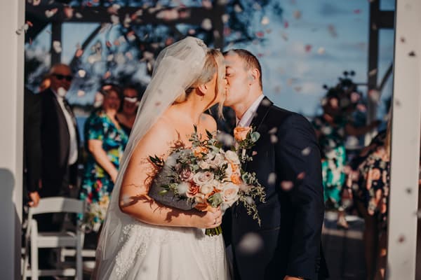Emily and Dylan kiss at the Sandstone Point Hotel — Pavilion ceremony stage as guests watch and flower petals fall around them.