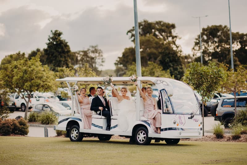 The bride Emily and groom Dylan sit together in a white open-sided vehicle at Sandstone Point Hotel, accompanied by three bridesmaids in pink dresses holding bouquets.