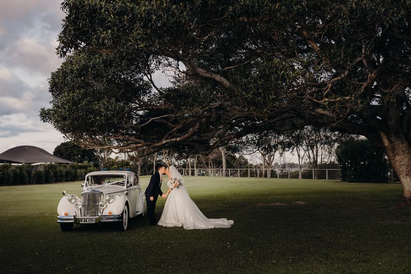 Emily the bride and Dylan the groom stand on grass near a vintage white car under a large tree at Sandstone Point Hotel, holding hands and facing each other.