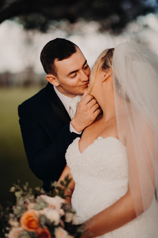 Emily the bride and Dylan the groom share a close moment during their couple portraits at Sandstone Point Hotel. Dylan gently holds Emily's chin as they lean in for a kiss. Emily wears a white strapless wedding gown and veil, holding a bouquet with peach and white flowers.