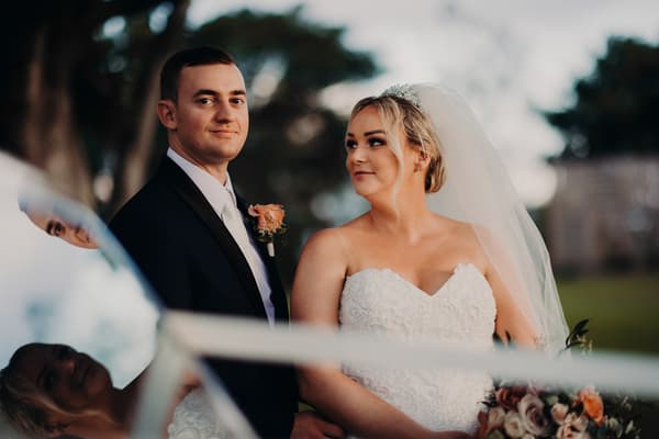 Emily in a white strapless wedding gown and veil holding a bouquet, and Dylan in a black suit with a white shirt and tie, pose together outdoors at Sandstone Point Hotel during their couple portraits session.
