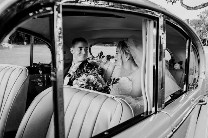 Emily the bride and Dylan the groom sit together in the backseat of a vintage car at Sandstone Point Hotel, with Emily holding a bouquet of flowers.