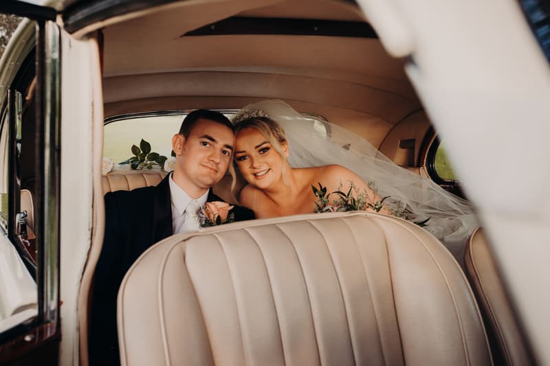 Emily the bride and Dylan the groom sit together in the back seat of a vintage car at Sandstone Point Hotel, with Emily holding a bouquet and wearing a veil and tiara.