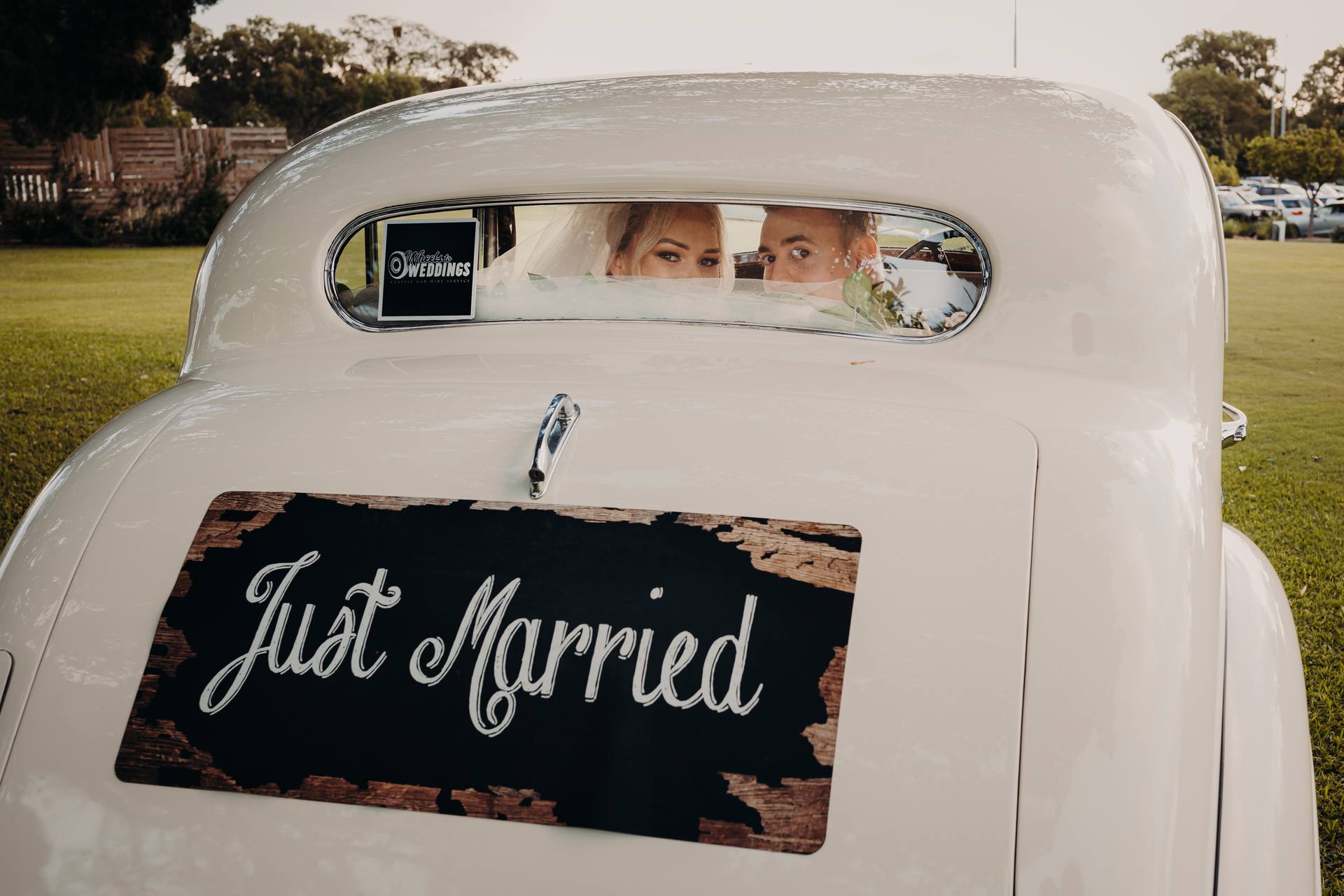 Bride Emily and groom Dylan sit inside a vintage white car with a 'Just Married' sign on the back at Sandstone Point Hotel, visible through the rear window.