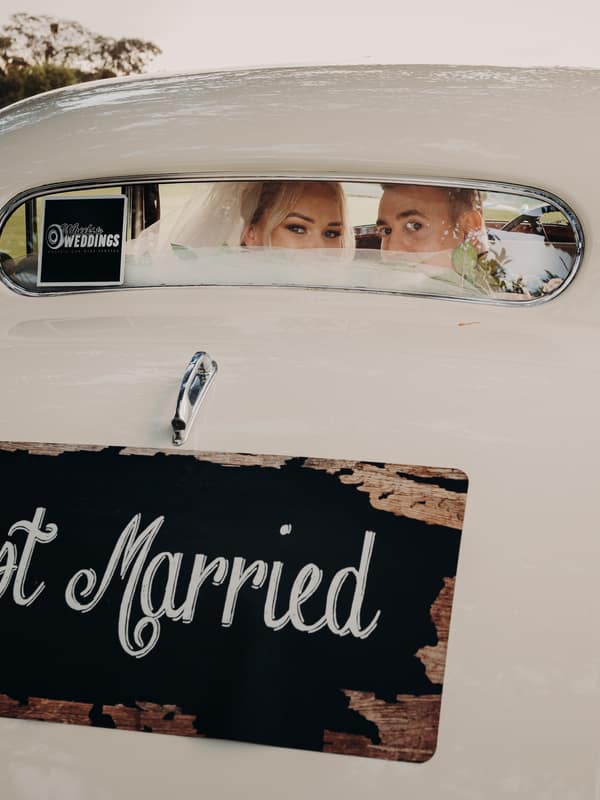 Bride Emily and groom Dylan sit inside a vintage white car with a 'Just Married' sign on the back at Sandstone Point Hotel, visible through the rear window.