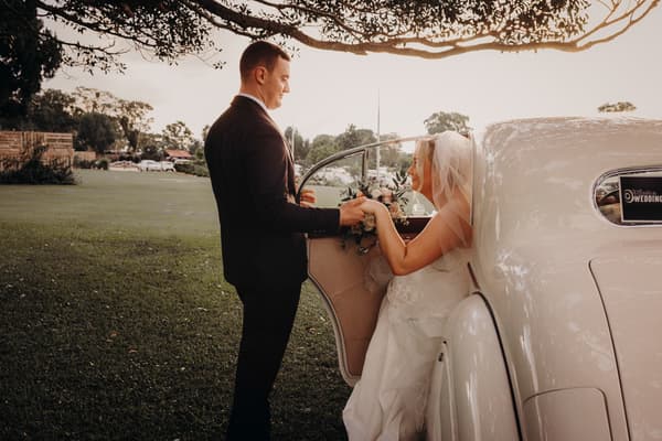 The groom stands outside a vintage white car holding hands with the bride, who is seated inside wearing a veil and holding a bouquet,. Best time of year to get married by She Said Yes.