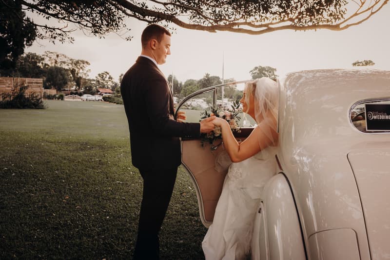 The groom stands outside a vintage white car holding hands with the bride, who is seated inside wearing a veil and holding a bouquet, at Sandstone Point Hotel.