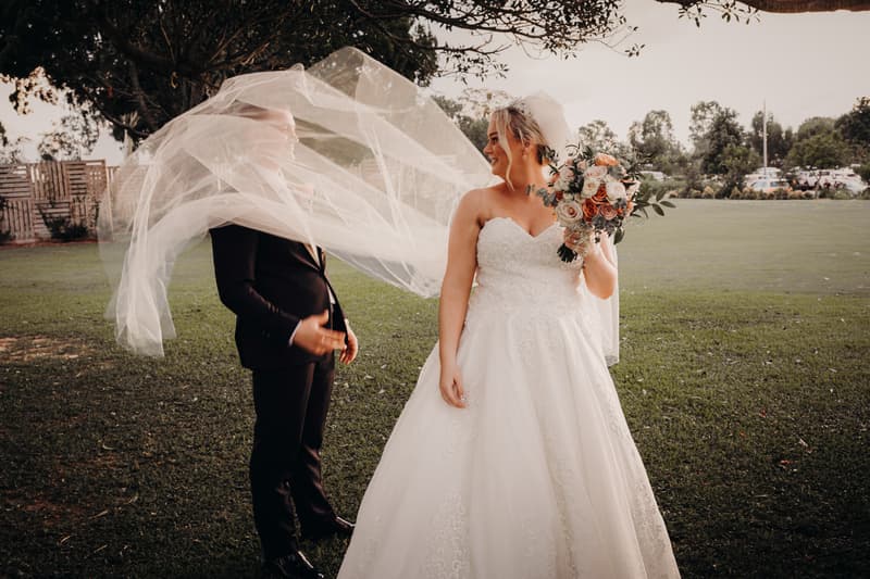 Emily the bride and Dylan the groom pose for couple portraits outdoors at Sandstone Point Hotel, with the bride holding a bouquet and her veil flowing over the groom.