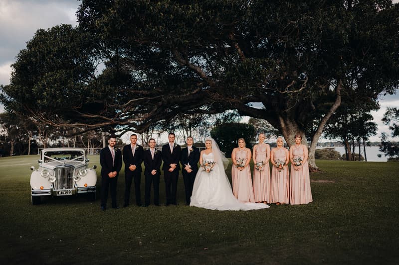 Emily the bride and Dylan the groom dance on a pier at Sandstone Point Hotel during sunset, with water and a bridge in the background.