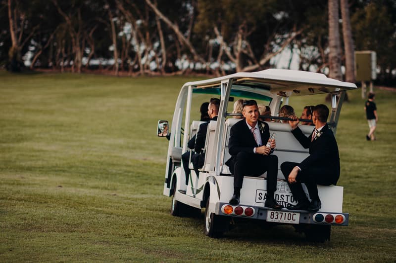 Several men in suits, likely groomsmen, sit on the back and inside a white golf cart on a grassy area at Sandstone Point Hotel, with trees in the background.