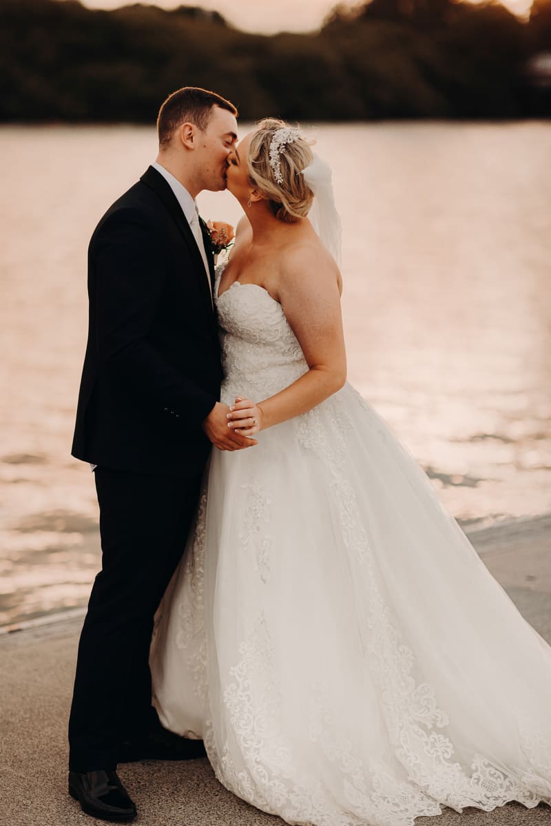 Emily the bride and Dylan the groom share a kiss while holding hands near the water. Wedding photography by She Said Yes.