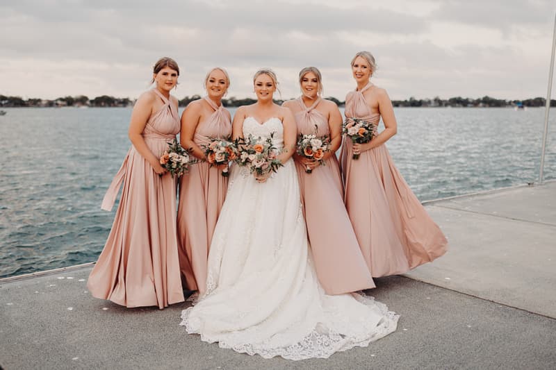 The bride Emily stands with four bridesmaids dressed in matching blush pink gowns, each holding bouquets, posing on a waterfront promenade at Sandstone Point Hotel.