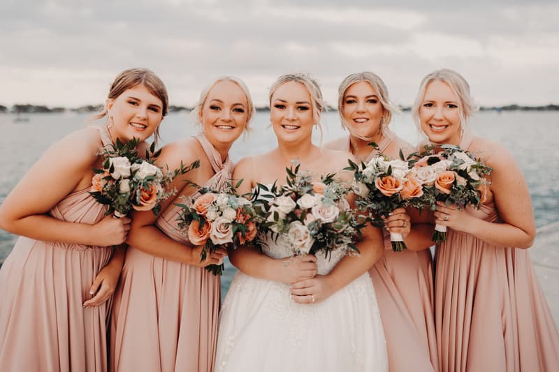 Emily the bride and Dylan the groom kiss at Sandstone Point Hotel while their bridal party, dressed in matching blush pink gowns and black suits, cheer and raise bouquets and hands by the waterfront.