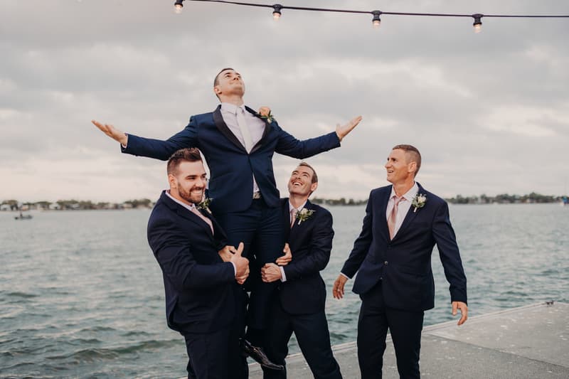 Dylan the groom is lifted by three groomsmen on a pier at Sandstone Point Hotel with water and cloudy sky in the background.