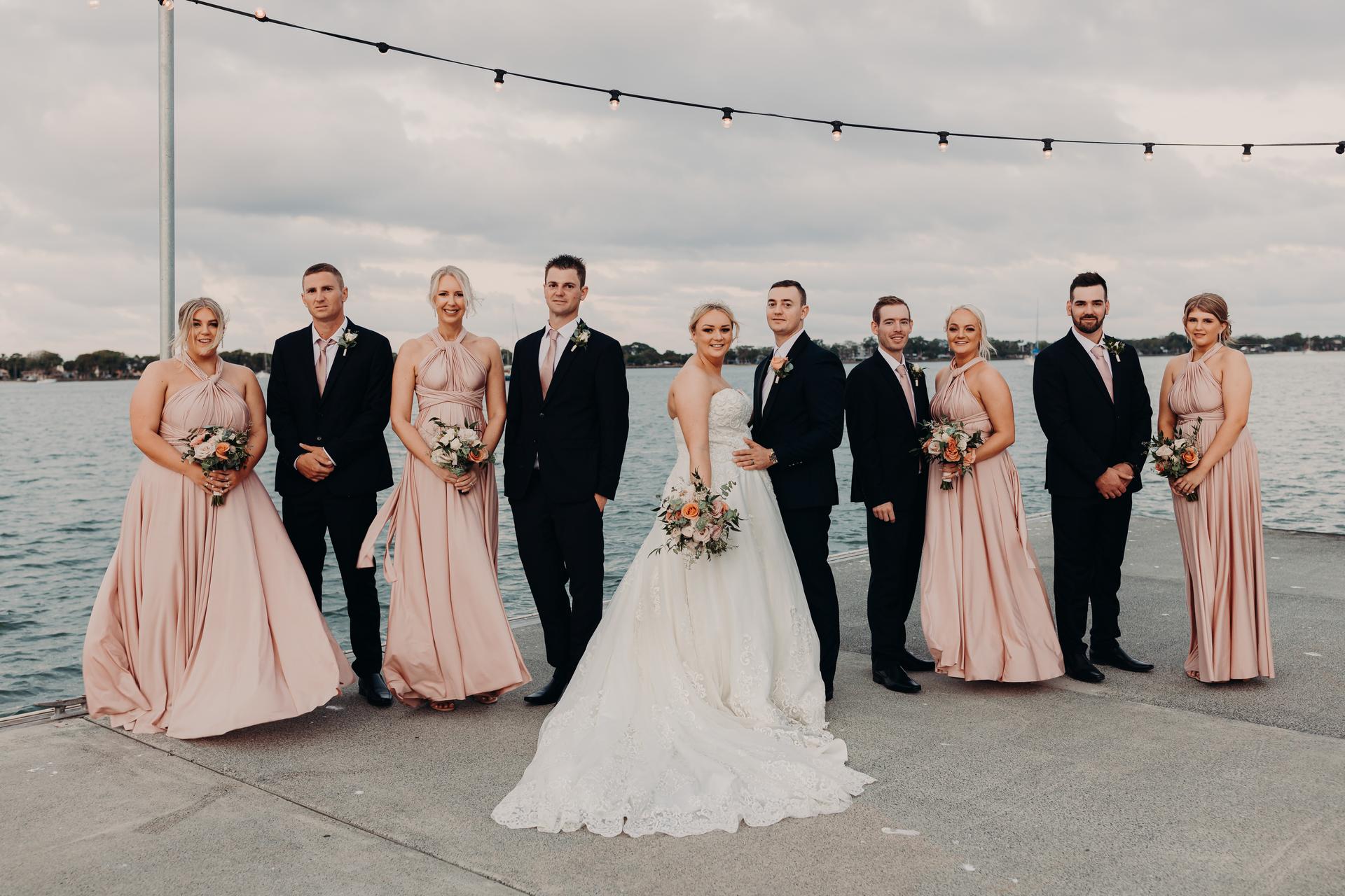 Emily the bride and Dylan the groom stand together with their bridal party at Sandstone Point Hotel, posing on a waterfront concrete pier under string lights. The bride wears a white gown and holds a bouquet, the groom and groomsmen wear black suits with pink ties, and the bridesmaids wear matching blush pink dresses holding bouquets.