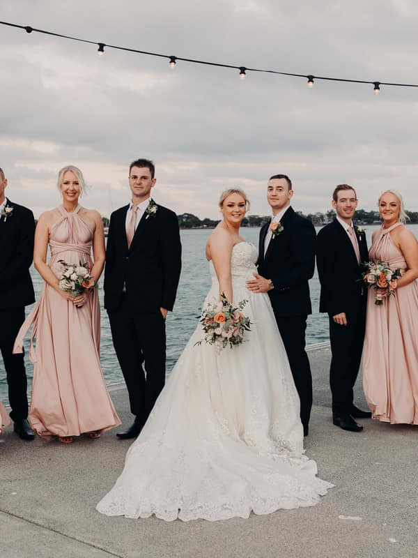 Emily the bride and Dylan the groom stand together with their bridal party at Sandstone Point Hotel, posing on a waterfront concrete pier under string lights. The bride wears a white gown and holds a bouquet, the groom and groomsmen wear black suits with pink ties, and the bridesmaids wear matching blush pink dresses holding bouquets.