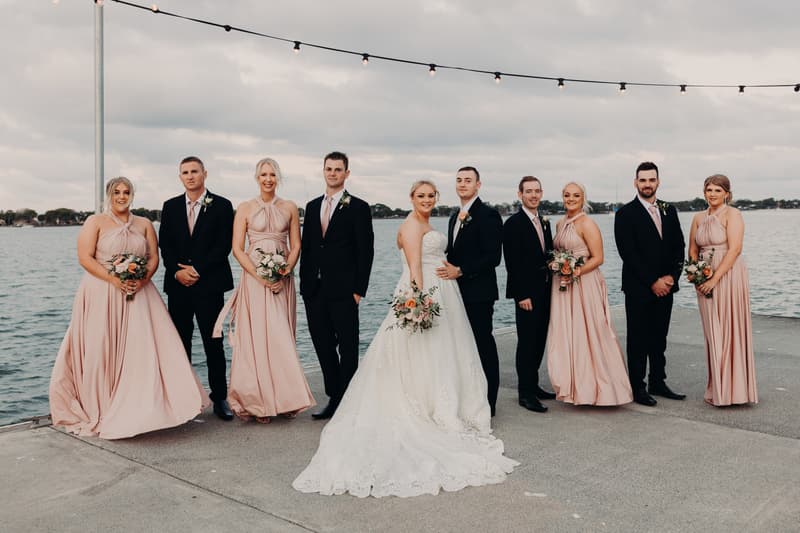 Emily the bride and Dylan the groom stand together with their bridal party at Sandstone Point Hotel, posing on a waterfront concrete pier under string lights. The bride wears a white gown and holds a bouquet, the groom and groomsmen wear black suits with pink ties, and the bridesmaids wear matching blush pink dresses holding bouquets.