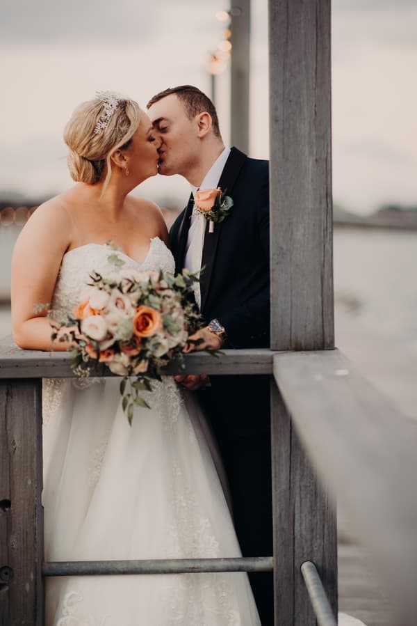 Bride Emily and groom Dylan kiss while standing on a wooden railing at Sandstone Point Hotel, with Emily holding a bouquet of flowers featuring peach and white roses.