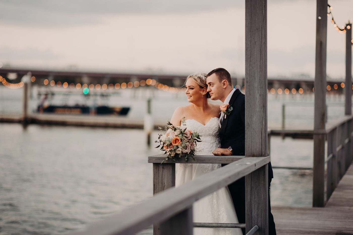Emily the bride and Dylan the groom stand together on a wooden pier, with Emily holding a bouquet of flowers and both looking out over the water. Winter wedding light photography Brisbane by She Said Yes.