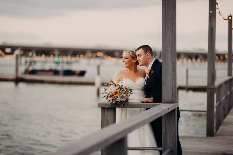Emily the bride and Dylan the groom stand together on a wooden pier at Sandstone Point Hotel, with Emily holding a bouquet of flowers and both looking out over the water.