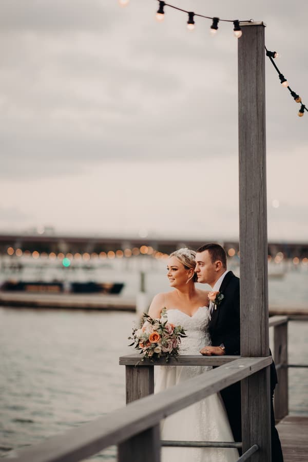 Emily the bride and Dylan the groom stand together on a wooden railing overlooking water at Sandstone Point Hotel, with Emily holding a bouquet of flowers and string lights overhead.