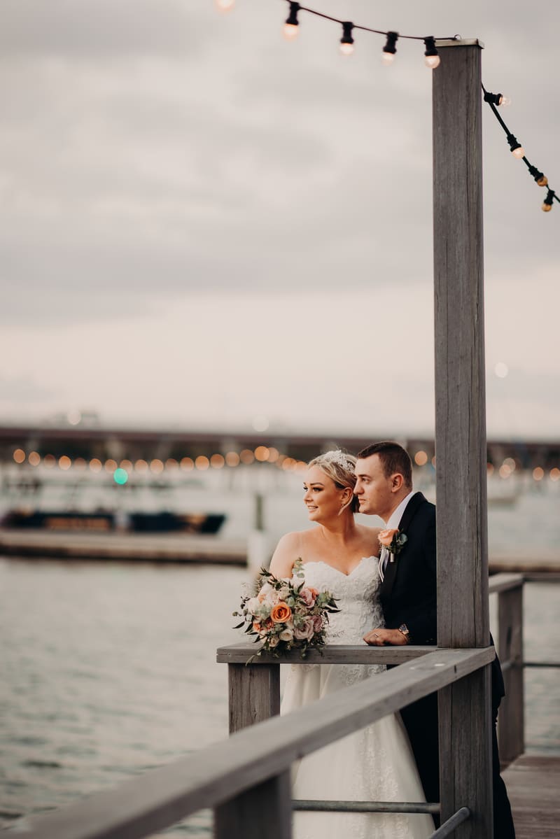 Emily the bride and Dylan the groom stand together on a wooden railing overlooking water, with Emily holding a bouquet of flowers and string lights overhead. Brisbane elopement photographer by She Said Yes.