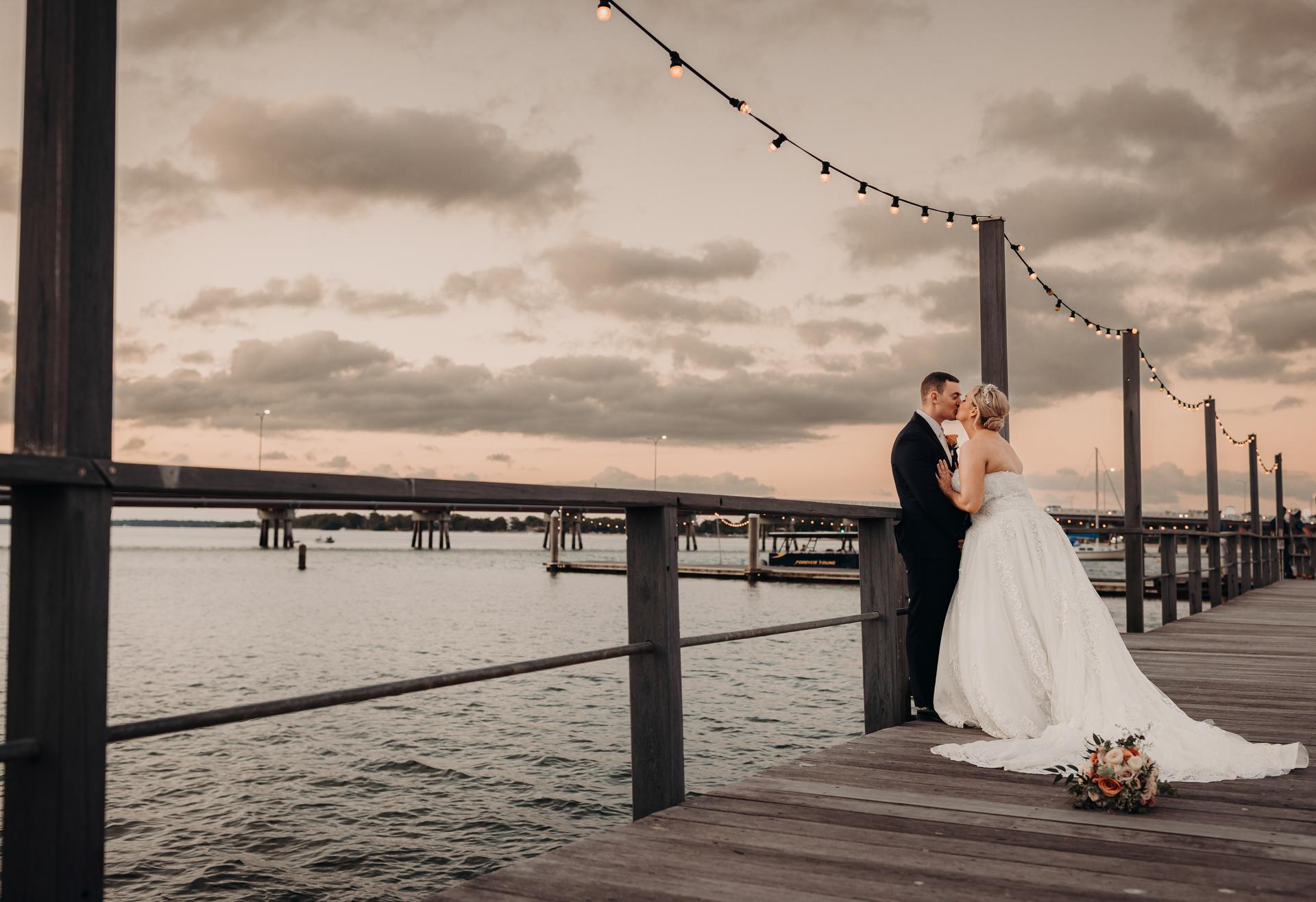 Bride Emily and groom Dylan share a kiss on a wooden pier at Sandstone Point Hotel during their couple portraits session at sunset, with string lights overhead and water in the background.