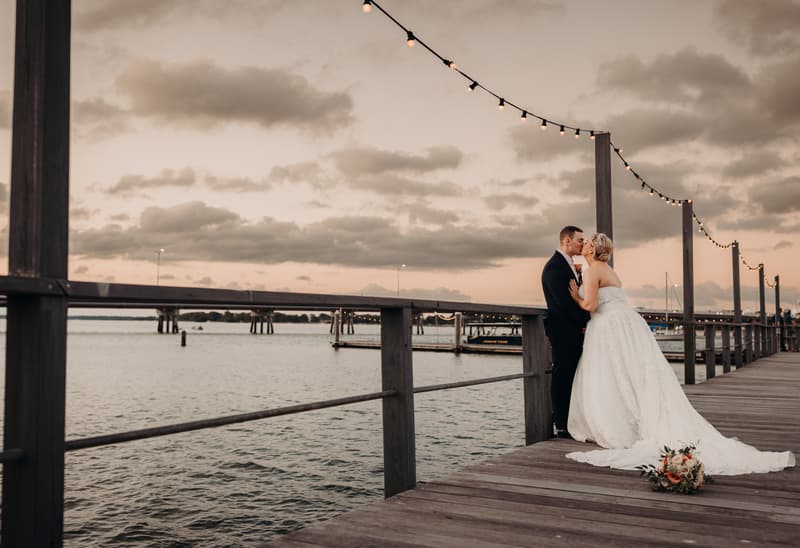 Bride Emily and groom Dylan share a kiss on a wooden pier at Sandstone Point Hotel during their couple portraits session at sunset, with string lights overhead and water in the background.