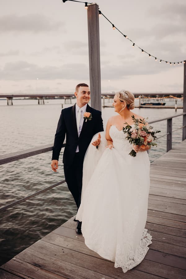 Emily the bride and Dylan the groom walk together on a wooden pier at Sandstone Point Hotel, with Emily holding a bouquet and Dylan holding part of her wedding dress. String lights hang overhead and a bridge is visible in the background over the water.
