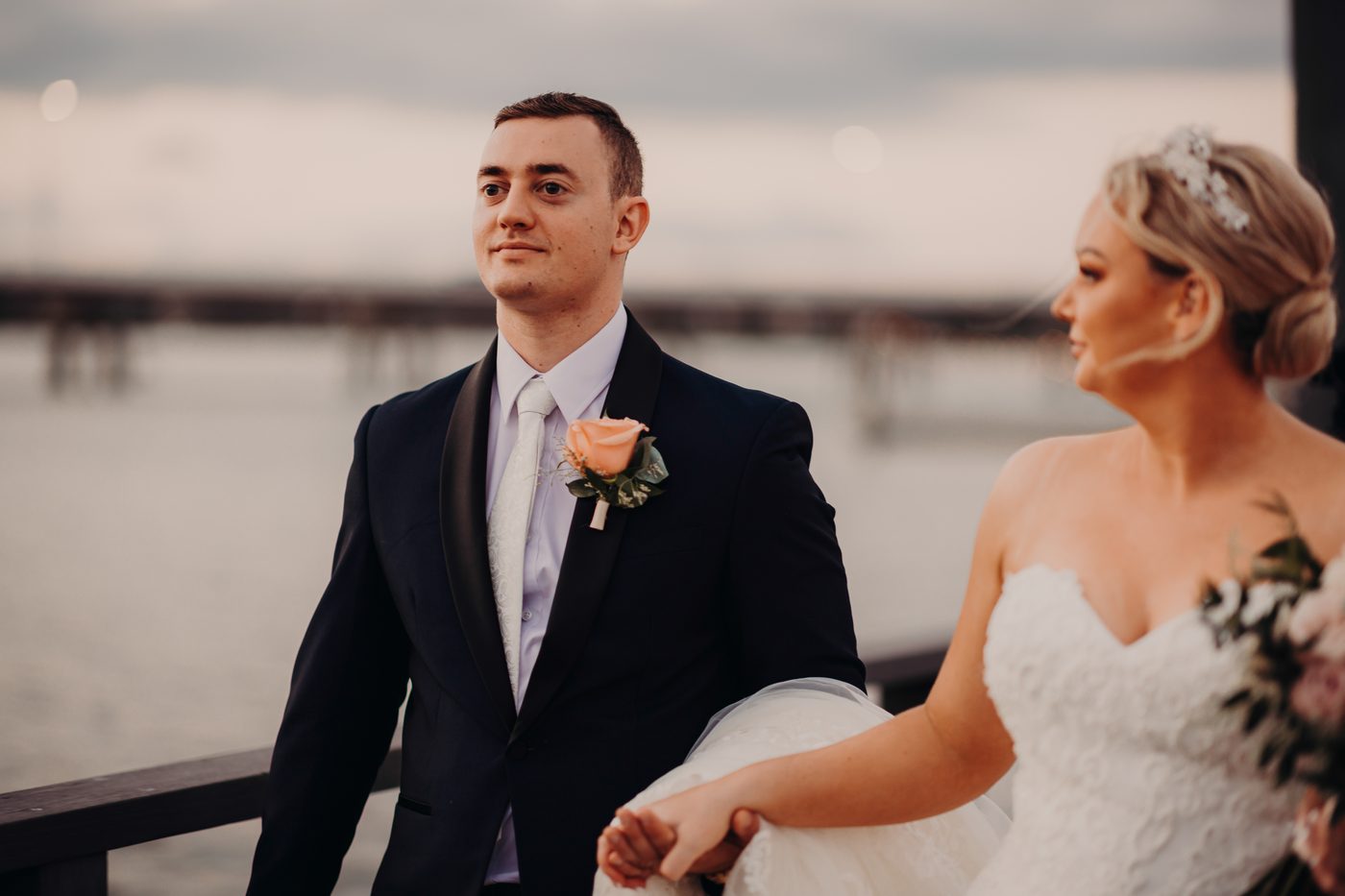 The groom Dylan in a dark suit with a peach boutonniere stands holding hands with the bride Emily in a strapless white wedding gown. Wedding photography pricing estimate by She Said Yes.