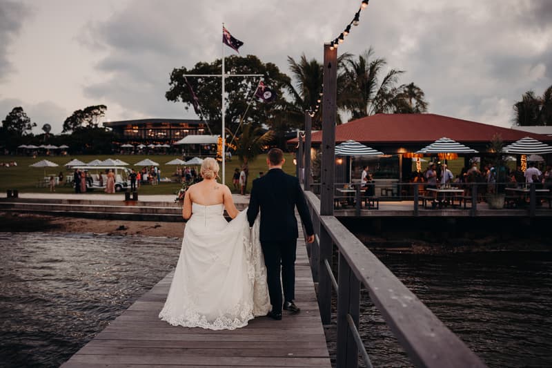The bride Emily and groom Dylan walk hand in hand along a wooden pier at Sandstone Point Hotel, with guests and umbrellas visible on the lawn and a building with a red roof to the right.