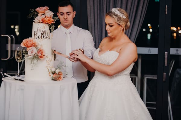 The bride Emily poses alone on the dance floor at the Sandstone Point Hotel — Pumicestone Room reception, holding out her white lace wedding gown with a chandelier and decorated head table in the background.