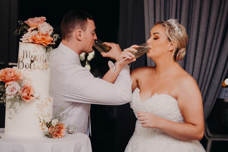 The bride Emily and groom Dylan share a dance on the wooden dance floor at Sandstone Point Hotel — Pumicestone Room, with a DJ booth and seated guests visible in the background.