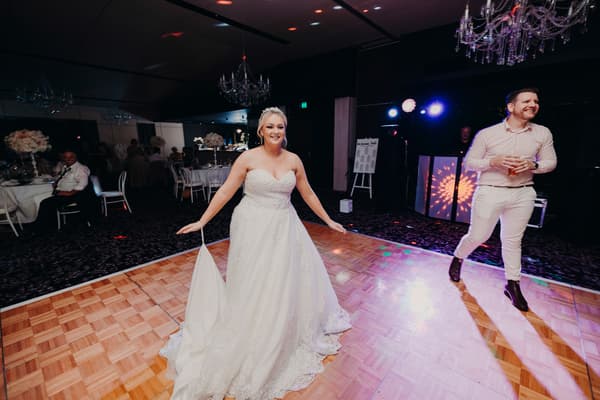 The bride and groom dance together at the reception in the Pumicestone Room at Sandstone Point Hotel, with guests and wedding party members visible in the background.