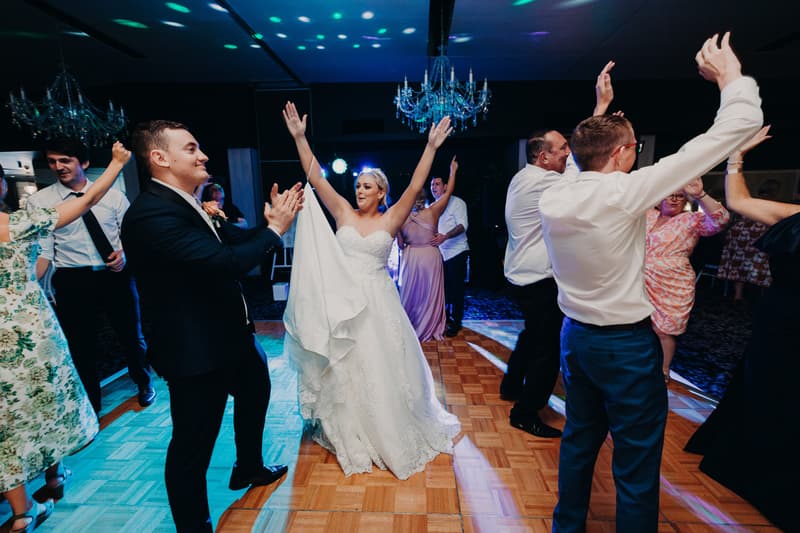 The bride Emily stands in the center wearing a white lace wedding gown and tiara, flanked by two men and two older women, all smiling, inside the Pumicestone Room at Sandstone Point Hotel.