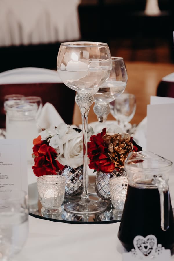 Table centerpiece at the Royal on the Park reception featuring floating candles in tall glass holders, red and white floral arrangements in silver vases, small votive candles, and a glass pitcher with a dark beverage.