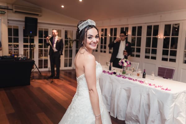 The bride Maryam in her wedding gown and tiara smiles at the camera inside Hillstone St Lucia — The Rosewood Room during the reception. A man in a suit is speaking into a microphone in the background, and another man is holding a drink near a table decorated with flowers and rose petals.