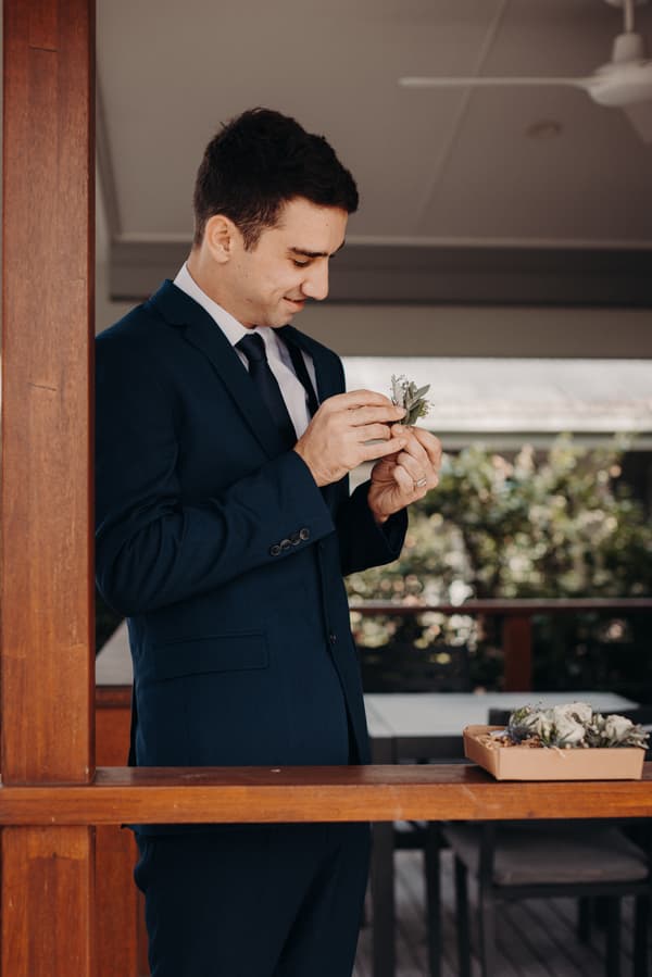 The groom Cameron stands at a wooden railing at Sandstone Point Hotel, adjusting a boutonniere from a box of floral arrangements on the table beside him.