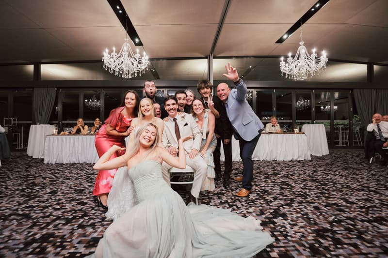 Morgan the bride and Joel the groom pose with a group of guests on the reception stage at Sandstone Point Hotel — The Pumicestone Room, with chandeliers and tables visible in the background.