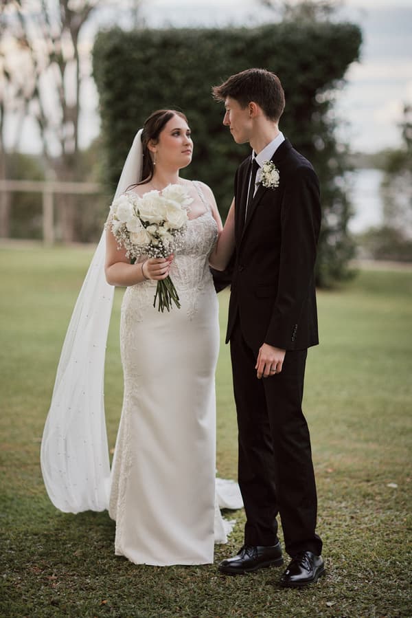 The bride Terion in a white wedding gown holding a bouquet of white flowers stands facing the groom Tyler in a black suit with a boutonniere at Sandstone Point Hotelallo outdoors on grass with greenery in the background.