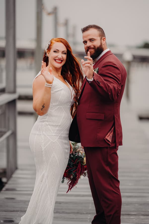 The bride and groom stand together on a wooden pier, both smiling and waving at the camera. The bride wears a white patterned gown and holds a bouquet, while the groom wears a burgundy suit with a boutonniere.