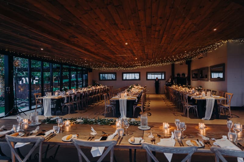 Reception tables set with glassware, cutlery, candles, and greenery at Preston Peak Winery Functions Centre, with fairy lights strung across the wooden ceiling and two people standing at the far end of the room.