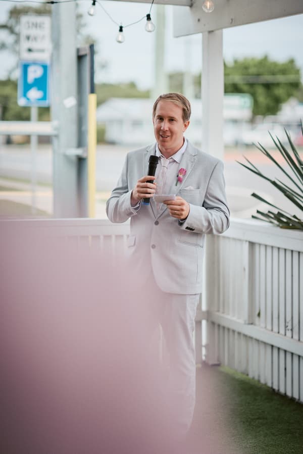 Jake, the groom, stands on a porch at White Horse Ranch holding a microphone and notes, delivering a speech during the wedding reception.